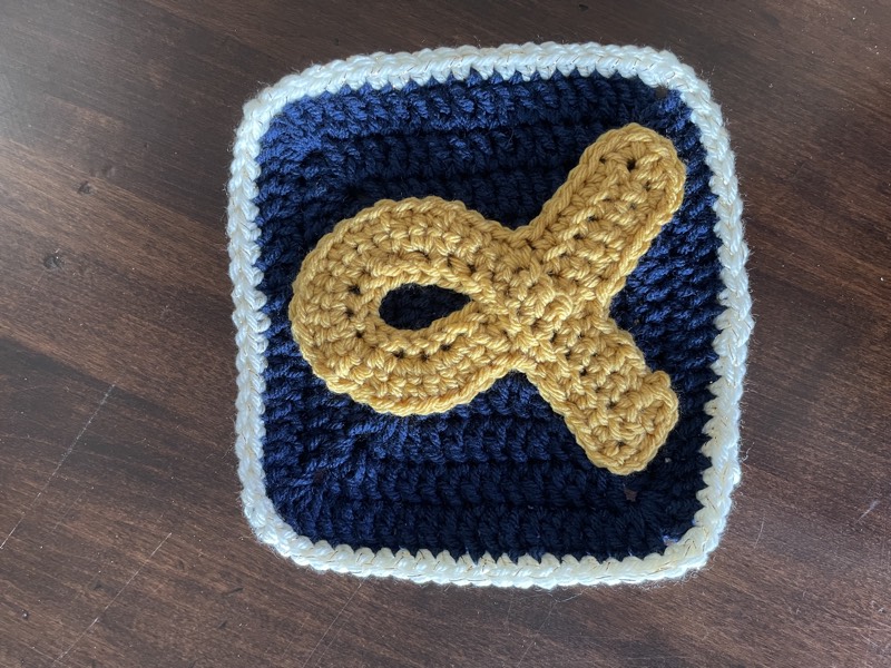 Crochet granny square with a gold childhood cancer awareness ribbon appliqué on a navy background